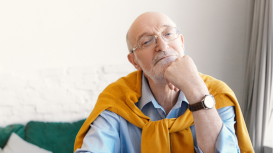 An older man with a sweater over his shoulders looks thoughtfully into the camera.