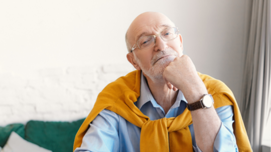 An older man with a sweater over his shoulders looks thoughtfully into the camera. 